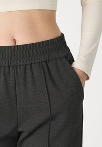 Pantalon de survêtement gris foncé avec une taille élastique et des poches latérales. Le tissu semble doux avec une texture subtile. Les détails incluent des plis.