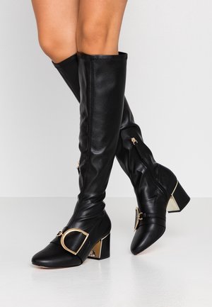 Bottes en cuir noires montantes jusqu'aux genoux avec talons carrés, boucles dorées et fermetures éclair latérales, portées par une personne aux jambes croisées.