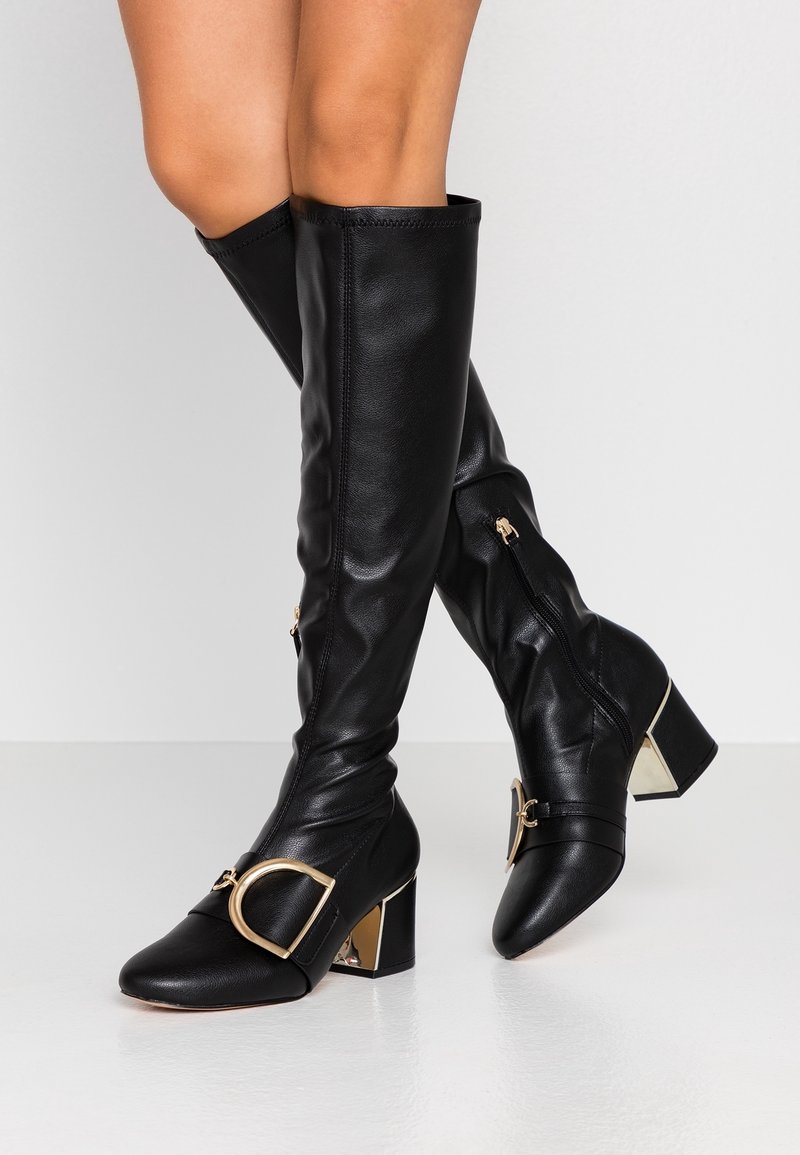 Bottes en cuir noires montantes jusqu'aux genoux avec talons carrés, boucles dorées et fermetures éclair latérales, portées par une personne aux jambes croisées.