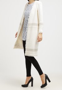 Pitkä, vaaleankreeminvärinen auki jätettävä neuletakki, jossa on teksturoitu kudontamalli ja vaaleita vaakaraidallisia yksityiskohtia. Päälle puettuna kuvioitu mekko ja mustat leggingsit.