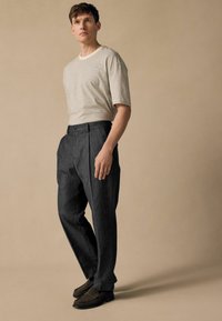 Camiseta de rayas claras con mangas cortas combinada con unos pantalones plisados de color gris oscuro. El modelo lleva mocasines marrón oscuro y está de pie frente a un fondo neutro.