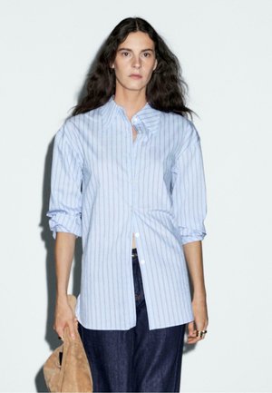 Lichtblauw overhemd met verticale witte strepen, voorzien van een puntige kraag en opgerolde mouwen, gedragen met donkerdenim broek.