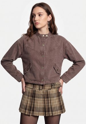 Young woman with brown hair wearing a brown cropped jacket and a pleated plaid mini skirt, standing with hands on hips looking to the side.