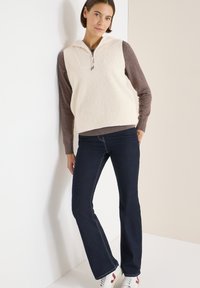 Fleecegefüllte Weste in Creme mit Kapuze, kombiniert mit einem braunen Langarmshirt und eng anliegenden dunklen Jeans. Akzente durch weiße Nähte und Sneakers.