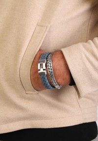 Leren gevlochten armband in lichtblauw naast een zilveren schakelarmband, gedragen om een pols, gedeeltelijk verborgen door een beige kledingstuk.