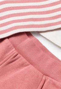 Rosa Jogginghosen mit einem strukturierten Bund, kombiniert mit einem weiß-rosa gestreiften Oberteil mit einem gerippten Saum. Beide Kleidungsstücke bestehen aus einem weichen Stoff.