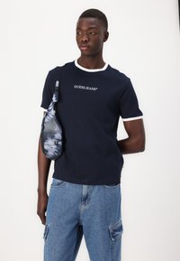 Camiseta azul marino con ribete blanco y logo "GUESS JEANS", combinada con pantalones cargo de mezclilla azul y una mochila de tirantes en tie-dye azul.