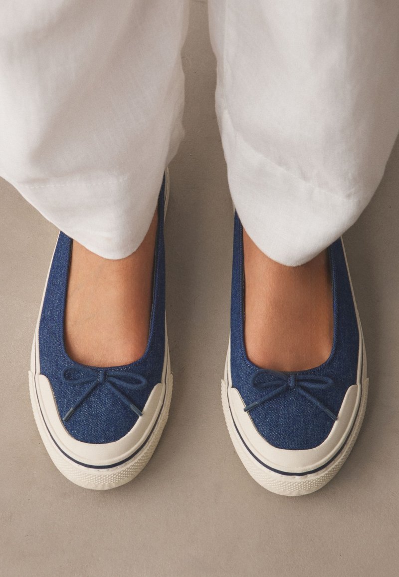 Pieds portant des chaussures bleu slip-on avec semelles en caoutchouc blanches et petits nœuds, associés à un pantalon blanc sur un fond de sol neutre.