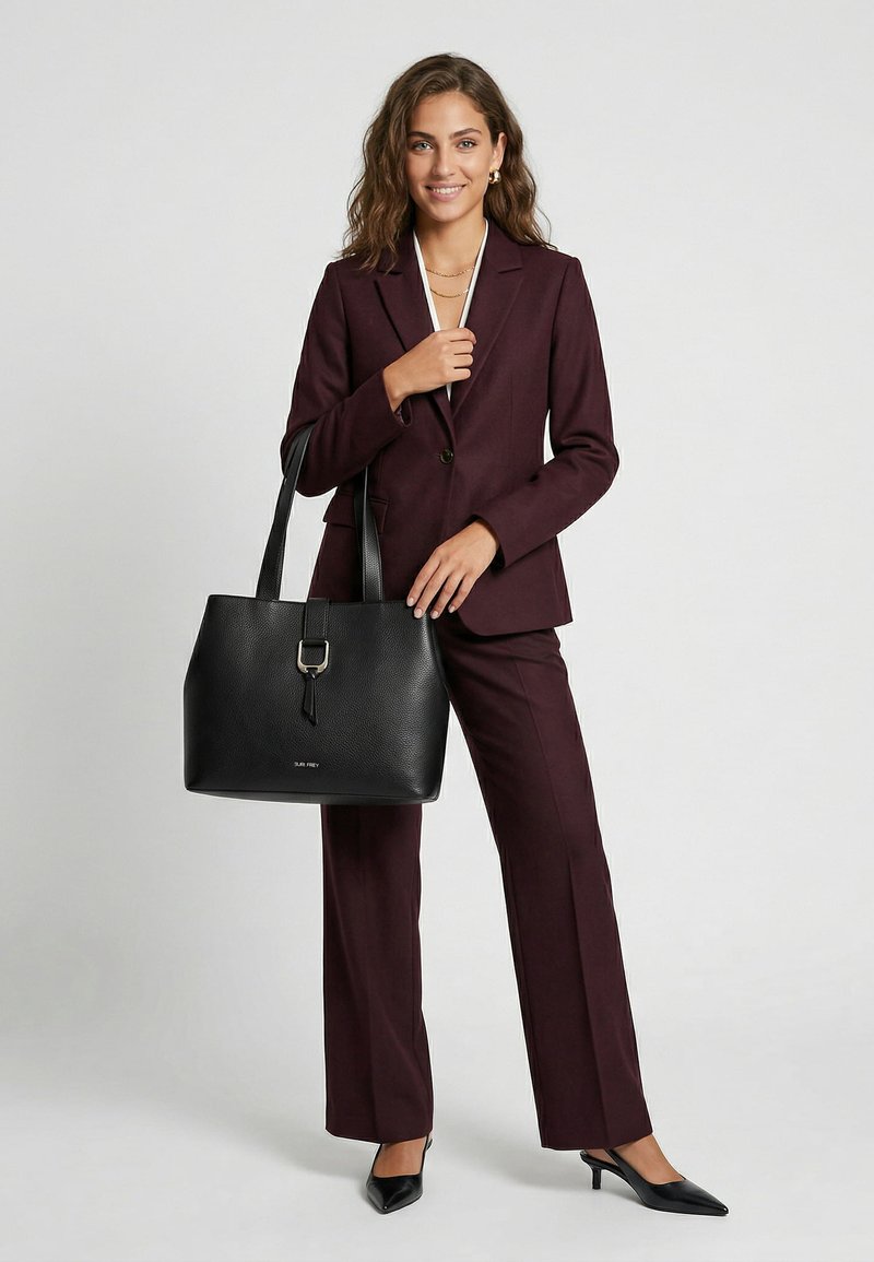 Donna in completo bordeaux scuro che tiene una grande borsa tote in pelle nera, indossa tacchi neri e gioielli in oro, sorridente su sfondo semplice.