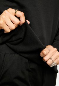 Melnas auduma tuvplāns ar roku, kas satver apakšu. Rokai ir vairāki gredzeni sudraba un zelta krāsā, kuros redzami dažādi dizaini un faktūras.