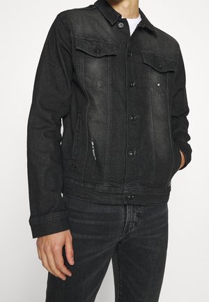 Homme portant une veste en jean noire délavée avec fermeture par boutons et un jean noir assorti sur un fond blanc uni.
