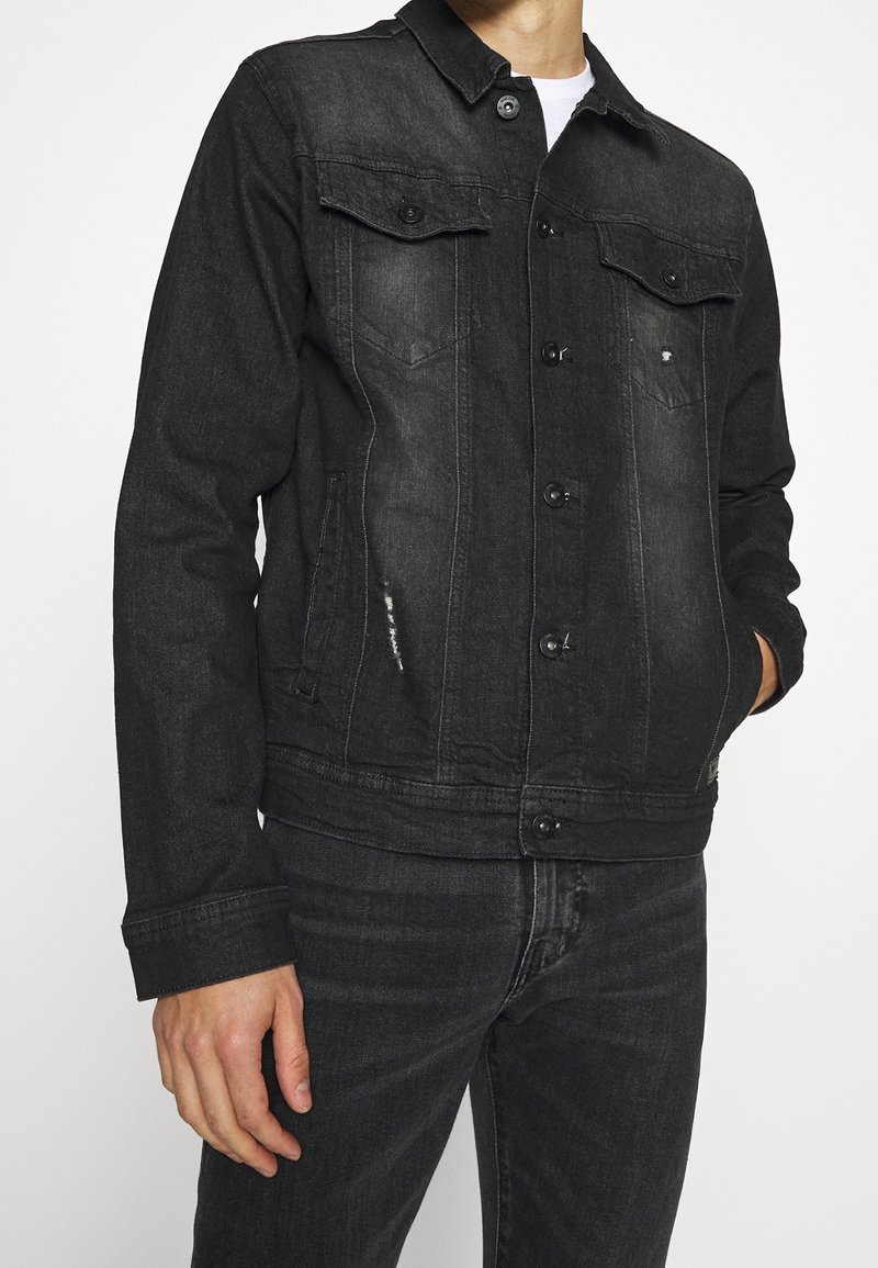 Homme portant une veste en jean noire délavée avec fermeture par boutons et un jean noir assorti sur un fond blanc uni.
