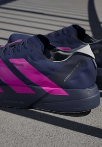 Chaussures de course bleu foncé avec des bandes rose vif et le texte "ADIZERO ADIOS PRO 4", posées sur une surface en béton avec des lignes d’ombre.