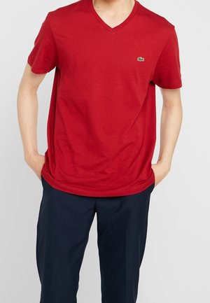 T-shirt en coton rouge avec un col en V, des manches courtes, et un logo brodé vert sur la poitrine gauche, associé à un pantalon bleu marine foncé.
