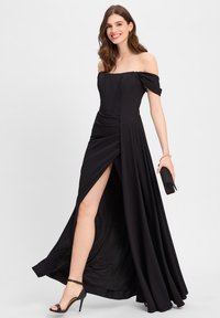 Schwarzes schulterfreies Kleid mit taillierter Oberseite, seitlichem Schlitz und fließendem Rock. Glattes Material, minimalistisches Design, kombiniert mit schwarzen Riemchen-Heels.