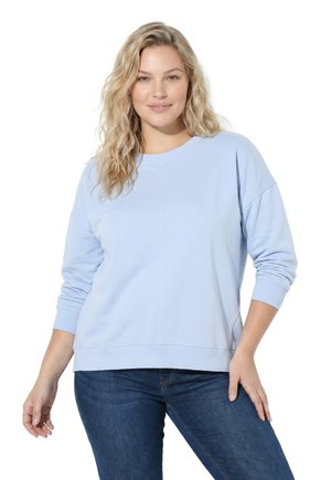 Kvinde med langt blondt hår iført en lyseblå sweatshirt og mørkeblå jeans, stående med den ene hånd på hoften mod en hvid baggrund.