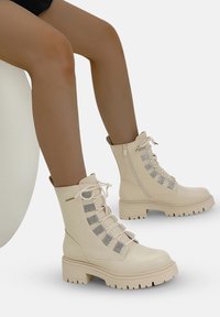 Bottes de combat beiges à lacets avec des panneaux en mesh, semelles épaisses et fermetures éclair latérales. Texture en cuir lisse et design de bout renforcé.