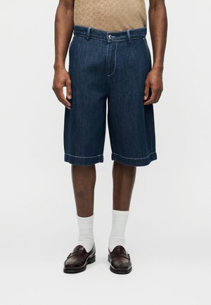 Muž nosiaci tmavomodré denimové šortky po kolená, biele členkové ponožky, hnedé mokasíny a béžovú košeľu s vzorom na jednoduchom pozadí.