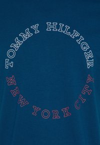 Modré bavlněné tričko s kulatým grafickým designem, na kterém je bíle napsáno "TOMMY HILFIGER" a červeně "NEW YORK CITY", umístěné uprostřed na přední straně.