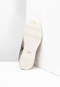 Beige Sneaker mit strukturierten Oberflächen, ausgestattet mit einer Gummisohle, markanter Branding und einem minimalistischen Design auf weißem Hintergrund.