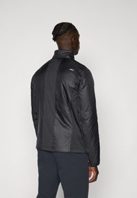 Black insulated jacket with a high collar, textured panels, and elastic cuffs. Features a logo on the upper back. Smooth fabric finish.