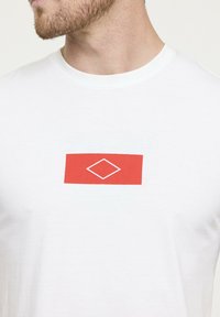 Gros plan sur une personne portant un T-shirt blanc à col rond avec un rectangle rouge et une forme de diamant blanche au centre de la poitrine.
