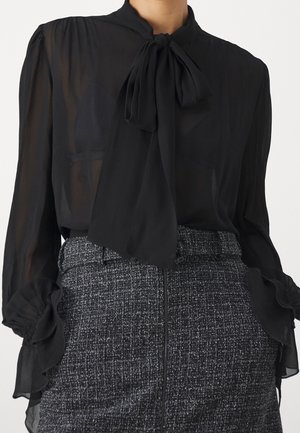 Zwarte doorschijnende blouse met grote strik bij de hals en geplooide manchetten, gecombineerd met een zwart-witte textuurrijke rok met hoge taille.