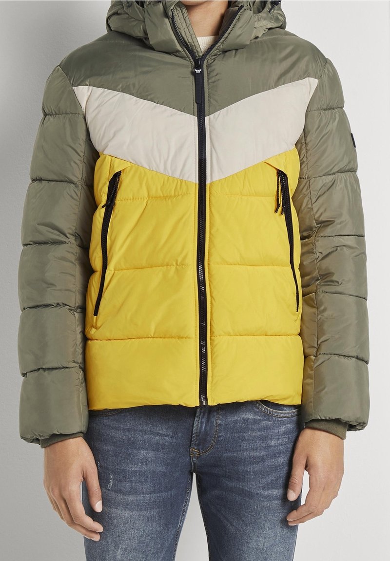 Uomo che indossa un giubbotto puffa colorato con maniche verde oliva, petto beige, torso giallo e tasche con zip, abbinato a jeans blu.