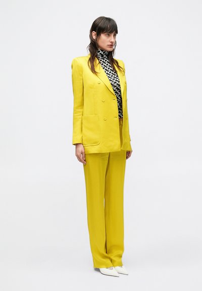 Blazer giallo su misura con design doppiopetto, abbinato a pantaloni coordinati. Sotto, un dolcevita con motivo in bianco e nero. Scarpe bianche.