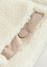 Valge fliiskangas, millel on pehme, tekstuuriga pind, beež kangariba ja ümmargune logo-nupp, millele on pressitud "BAKER".