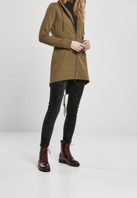 Olivově zelená mikina na zip s kapucí na šňůrku a předními kapsami, spárovaná s černými texturovanými legínami a lesklými burgundskými šněrovacími botami.