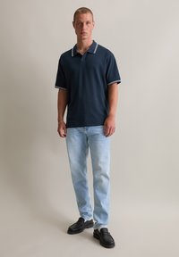 Granatowa koszulka polo z białym paskiem na kołnierzyku, zestawiona z jasnoniebieskimi dżinsami i czarnymi butami wsuwanymi, stojąca na neutralnym tle.