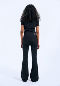 Femme aux cheveux bouclés, portant un t-shirt noir ajusté et un pantalon noir évasé taille haute, debout de dos sur un fond blanc.