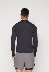 Μακρυμάνικο αθλητικό πουκάμισο σε στενή εφαρμογή, μαύρο, με raglan μανίκια, συνδυασμένο με γκρι σορτς. Το ύφασμα είναι λείο και ελαστικό, σχεδιασμένο για άνεση.