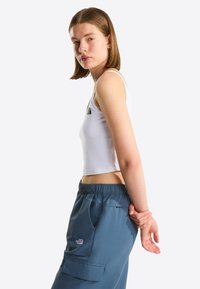 Fiatal nő fehér, rövidített fazonú felsőben és kék cargo nadrágban, oldalra fordulva, kezeit a háta mögött összekulcsolva egy egyszínű háttér előtt áll.