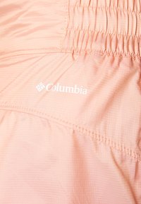 Casaco Columbia rosa claro com faixa elástica franzida na cintura; leve, de textura suave; apresenta um logotipo branco na parte inferior.