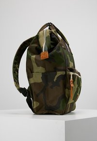 Reppu, jossa on vihreä ja ruskea maastokuvio, edessä vetoketjullinen tasku ruskealla nahkahihnalla ja pehmustetut olkahihnat.