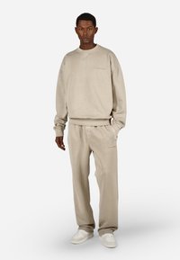 Muž oblečený v béžových oversize mikinách a ladiacich teplákoch s bielymi teniskami, stojaci na jednotnom svetlom pozadí.