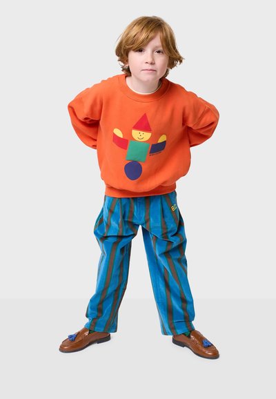 Sudadera naranja con un diseño colorido de figura geométrica; pantalones a rayas azules y marrones; zapatos mocasines marrones con borlas.