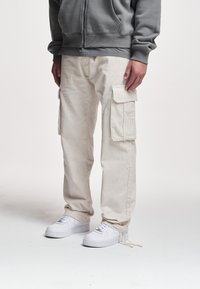 Beige cargo bukser med ribbet tekstur, udstyret med store sidelommer, parret med en grå lynlås hoodie og hvide sneakers.