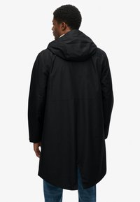 Zwarte waterdichte jas met een capuchon, met een langere achterkant, lange mouwen en een gladde textuur. Subtiele branding op de achterkant.