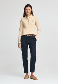 Polo beige a maniche lunghe con colletto, cinque bottoni e logo; abbinato a pantaloni blu scuro e mocassini beige. Sfondo semplice.
