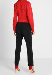 Veste en daim rouge associée à un pantalon noir, avec des talons rouges hauts. La veste a une coupe ajustée et une texture lisse.