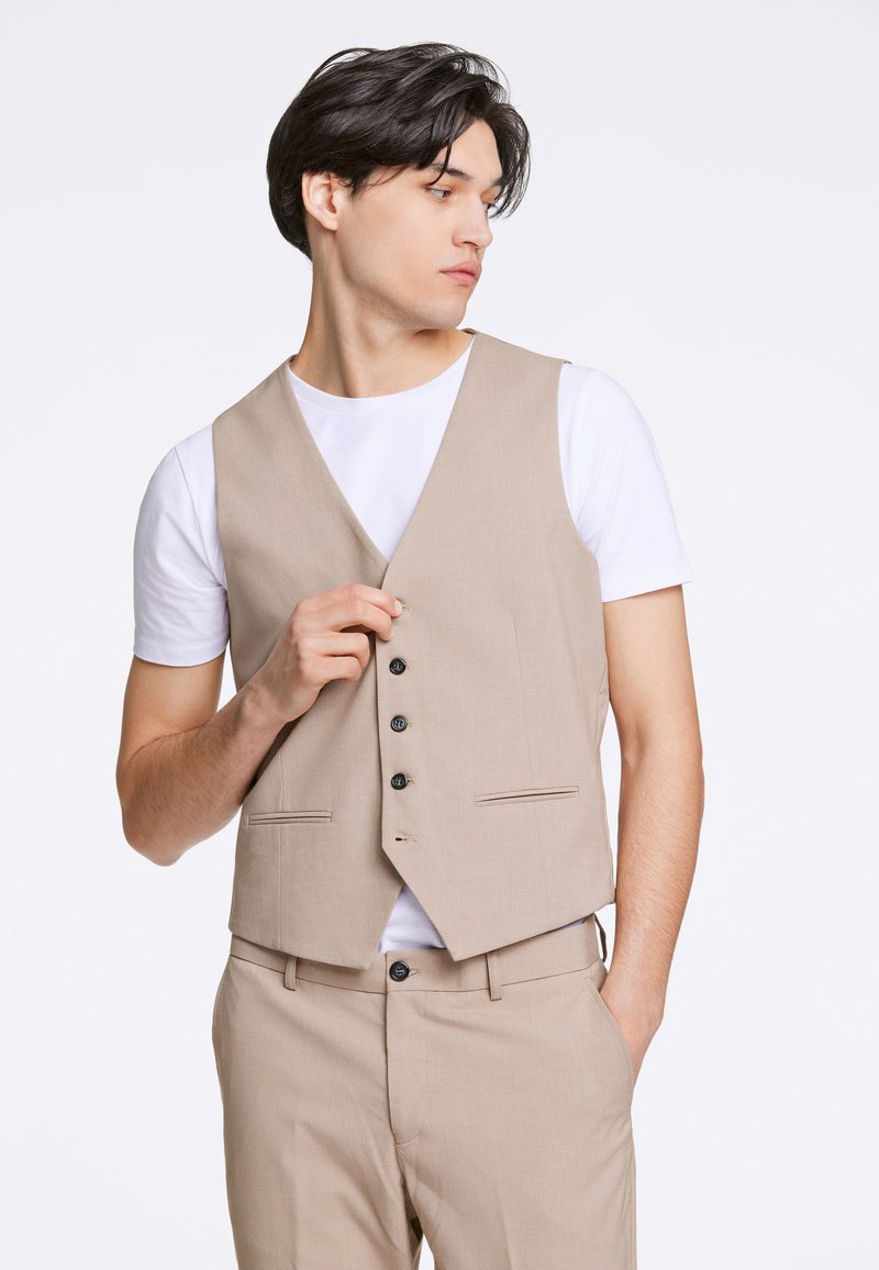 Mladi moški zapenja bež telovnik čez belo majico s kratkimi rokavi, oblečen v ujemajoče bež hlače, gleda stran proti enobarvnemu ozadju.