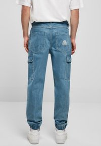 Denim cargo bukser i lyseblå med elastiske manchetter, to baglommer, cargolommer på siden og et hvidt logo broderet på bagsiden.