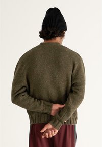 Olive zielony sweter z dzianiny z prążkowanym wykończeniem i długimi rękawami. Zawiera pomarańczowe drobinki. W zestawie z czarną prążkowaną czapką.