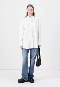Chemise blanche à manches longues avec un petit logo, associée à un jean large bleu clair et des chaussures à plateforme noires, tenant un sac à main noir.