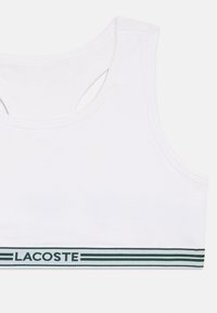 T-shirt branca sem mangas com decote redondo, costas recortadas e uma faixa verde na parte inferior com o texto "LACOSTE". Confeccionada em um tecido de algodão suave.