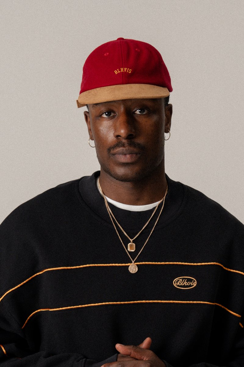 Gorra roja con visera beige, que presenta el texto "BLKVIS." Se lleva con un sudadera negra, acentuada por rayas naranjas y múltiples collares de oro en capas.