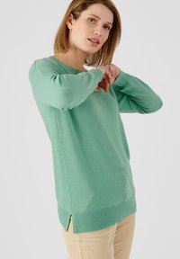 Jersey verde claro hecho de material suave, con cuello redondo, mangas largas y aberturas laterales en el dobladillo para facilitar el movimiento.